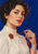 Close portrait of a woman with short dark curls holding a crimson ribbon, wearing an ivory blouse and red floral brooch against a blue background.