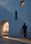Small cyclist beside a vivid red bicycle at a pale-blue textured wall with three openings, warm-lit windows, and long pavement shadows.