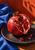 Opened pomegranate with glossy ruby seeds on a dark ceramic plate, with cobalt-blue cloth and terracotta wall behind.