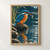 Blue-and-rust kingfisher perched on a rock holding a small fish above a dark river ripple, with reeds and falling droplets.; frame shown for styling only