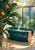 Deep teal freestanding bathtub with steam and a coral towel in a bright glass conservatory, framed by broad green foliage and rain-streaked windows.