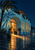 A blue-hour seaside pavilion with glowing amber windows, an arched entrance, palm tree and reflections on wet paving beside water.