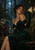Adult woman in a dark emerald gown seated in a moonlit glasshouse, looking toward a glowing moth near her raised gloved hand.