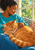 A ginger cat curled asleep in an adult woman's lap by a bright window, with turquoise clothing and patterned cushions.