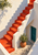 Red-orange zigzag staircase in a sunlit plaster courtyard, with teal door, terracotta planter, and leafy branches.