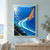 Small white sailboat with a vivid orange triangular sail cutting across a blue bay and leaving a broad curling wake, with wooded headlands and distant mountains.; frame shown for styling only