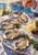 Hand lifting an oyster with a fork above crushed ice and opened shells, with lemon, dark sauce, a glass and blue sea beyond.