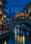 Vintage-style streetcar crossing a stone arch bridge over a night-time city canal, with glowing windows, water reflections, and a cyclist below.
