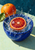 Halved blood orange in a cobalt-blue bowl surrounded by a crown of water, with a whole orange and green leaf behind it.