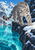 Spotted snow leopard on a snowy rocky ledge above a turquoise glacial pool and jagged mountains.