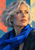 Close portrait of a silver-haired woman wearing a cobalt scarf against a geometric coral, teal and pale yellow background.