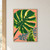 Oversized emerald monstera leaf backlit with golden openings and a curled young leaf against coral ground.; frame shown for styling only