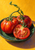 Three red tomatoes, one cut open to show its seed chambers, rest on a dark green dish against a yellow-orange background.