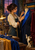 A tailor pins a cobalt lapel onto a dress form while a calico cat reaches for thread in a warmly lit sewing studio.