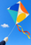 Multicolored geometric kite with sweeping tails against blue sky, held by a gloved hand at the lower left.