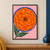 A large orange marigold-like flower viewed from above, with a dotted dark center, blue circular ring and pointed green leaves on pink.; frame shown for styling only