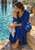 Adult woman in a deep-blue robe seated on a stone pool ledge and squeezing water from a towel beside turquoise water.