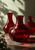 Three glossy red glass carafes, one large vessel in front of two narrower ones, against a pale wall with leafy shadows and small title text.
