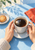 Two hands in cream sleeves holding a white cup of dark coffee over a saucer, beside a biscuit, glass vase and red-orange napkin.