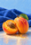 Cut apricot exposing orange flesh and a brown stone beside a whole apricot with a green leaf, arranged against cobalt-blue cloth.