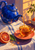 Cobalt-blue teapot pouring amber tea into an amber cup on a coral saucer, with a sliced blood orange, green leaf, striped cloth, and warm window light.