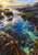 Blue-green tidepool among dark coastal rocks with receding white foam and a small orange sea star beneath a warm sunset sky.