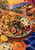 Ceramic platter of flatbread, olives, figs, white cheese, and pomegranate on a terracotta table, with a hand offering olives and a sunset coast behind.