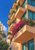 A pale terracotta apartment building with repeating planted balconies and cascading magenta flowers against blue sky and neighboring buildings.