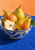 Three whole pears and a cut pear half in a blue-and-white patterned bowl on a blue table against an apricot wall.