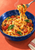 Fork lifting tagliatelle from a blue bowl with tomato pieces and basil on a coral tabletop.