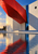 Modern pale-concrete pavilion with an angular red canopy, blue-glass panel, round wall opening, and reflecting pool.