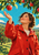 An adult woman in a bright red coat looks up and reaches toward a red apple beneath leafy branches, with orchard rows and low hills behind her.