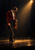 Full-figure violinist in a crimson jacket bows their head while lowering a violin beneath a warm spotlight.
