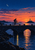 Small silhouetted cyclist with an amber light on a stone bridge, with a lighthouse and sunset beyond.