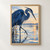 Blue-gray heron lowering its bill toward shallow dawn water, with reeds and a warm sunrise reflection.; frame shown for styling only