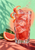 A tall faceted glass of pink-red grapefruit soda with ice, curled peel and bubbles, beside a grapefruit wedge and leaves.