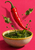 Glossy red chili pepper above a ceramic bowl of green herb sauce, with droplets and leaves against a warm pink background.