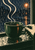 Graphic poster-style scene of a hand holding a dark-green teacup on a rain-speckled windowsill, with pale steam and an amber streetlamp outside.