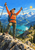 An orange-jacketed hiker raises two trekking poles on a rocky overlook above a turquoise alpine lake and layered mountains.