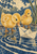 Two yellow ducklings stand in indigo ripples beside a cream folded paper boat, with a green reed at the right.