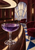 Faceted coupe of violet liquid on a curved dark wood counter in an elegant lounge with burgundy chairs and a fan-shaped window.