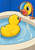 Yellow rubber duck in an ivory basin facing a coral-rimmed magnifying mirror that shows an enlarged eye and beak, against a cobalt-blue tiled wall.