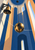 Reflective brass pendulum bob hanging beside a brass bell in a tall shaft of cobalt, cream, and apricot architectural planes.