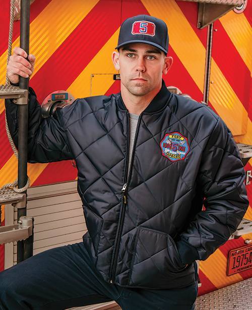 Firefighter in jacket and custom-embroidered cap holding on to the back of a firetruck