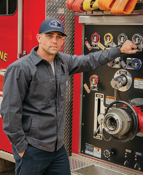 Firefighter with a custom firefighter hat leaned against a pump panel of a firetruck