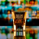 Wholesale engraved 2.25 oz. V Shot Glass customized with Dueling Grounds Distillers logo on a bokeh background
