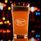 Wholesale engraved 20 oz. English Nonic Pub Glass customized with Cabbage Shed logo on a bokeh background