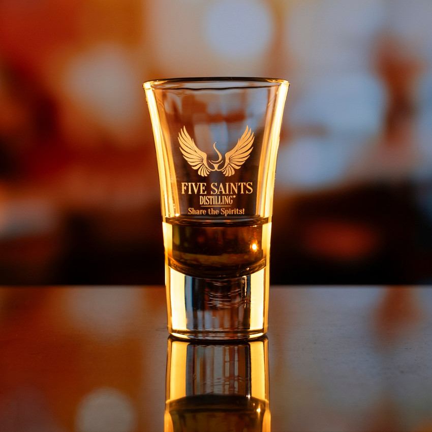 Wholesale engraved 2 oz. Moon Shot Glass customized with Five Saints Distilling logo on a bokeh background