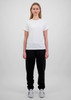 A woman in a white t-shirt and black track pants stands against a plain background. The pants have a branding logo.
