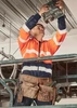 A worker in an orange and navy hi-vis full zip hoodie uses a tool while standing on a scaffold, wearing a hard hat.
